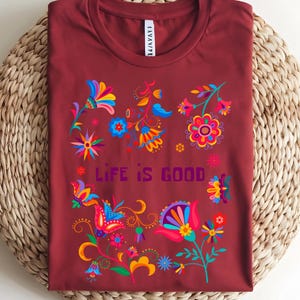 May include: A maroon t-shirt with a colorful floral design and the text "LIFE IS GOOD". The flowers are in shades of blue, yellow, pink, and orange. The shirt is folded on a woven surface.
