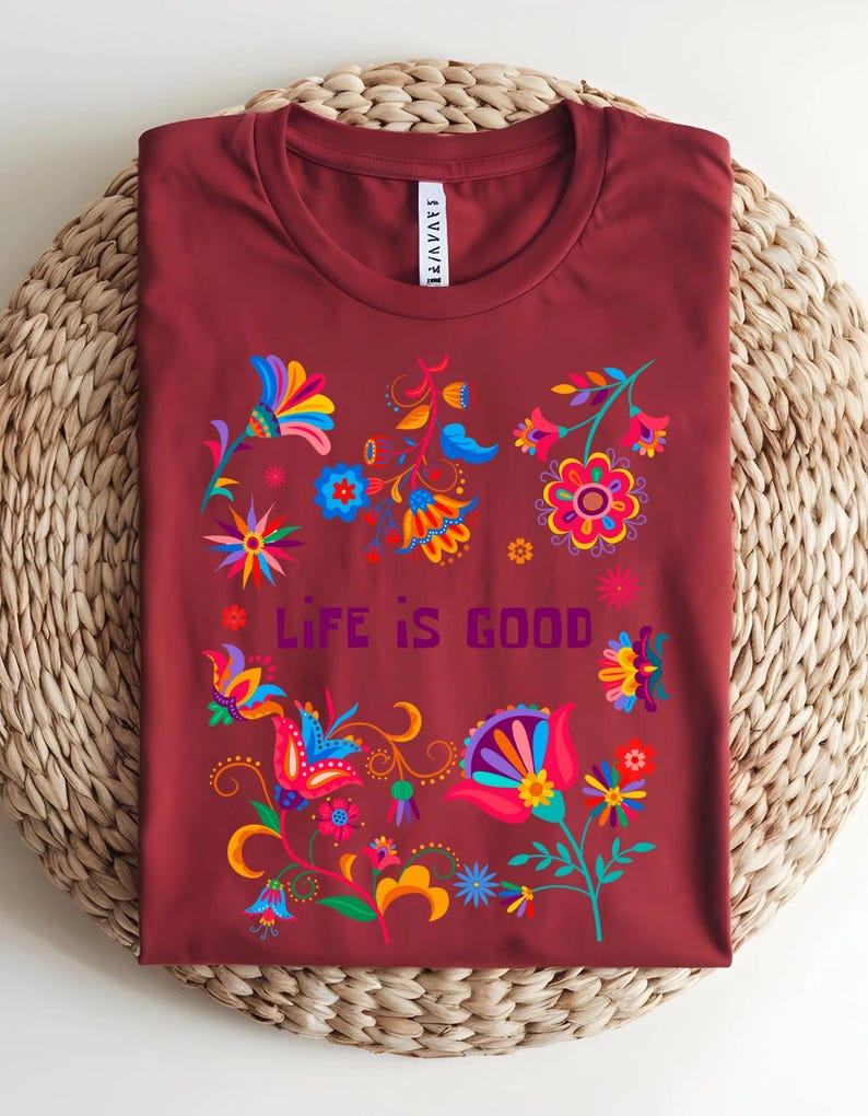 May include: A maroon t-shirt with a colorful floral design and the text "LIFE IS GOOD". The flowers are in shades of blue, yellow, pink, and orange. The shirt is folded on a woven surface.