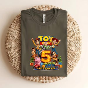 May include: A dark olive green t-shirt featuring a colourful Toy Story graphic with Woody, Buzz Lightyear, Jessie, Bullseye, and other characters surrounding a large yellow number 5. The text reads Toy Story, Andy's Room 2026.