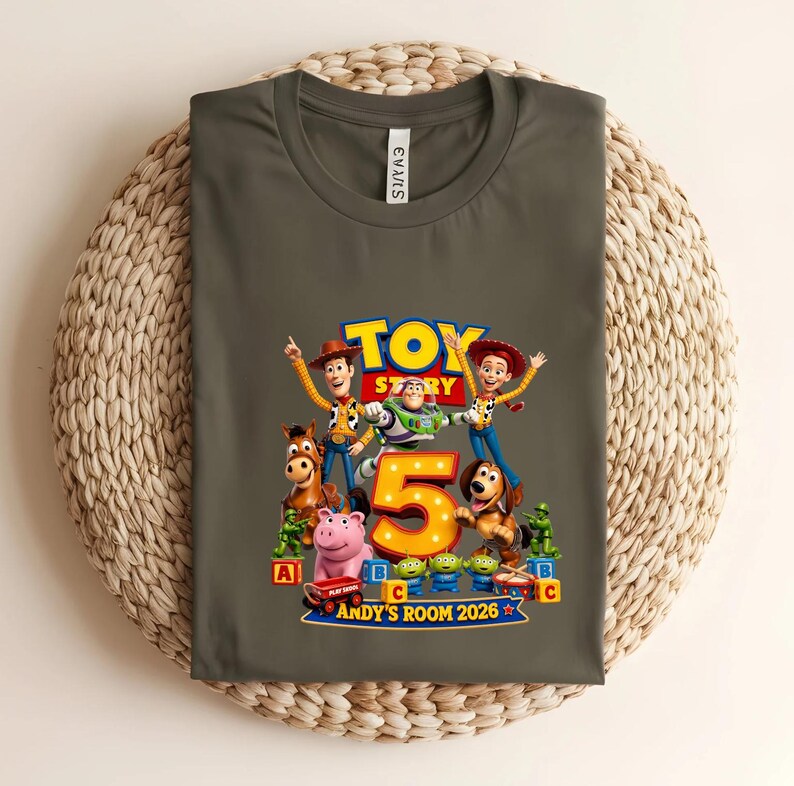May include: A dark olive green t-shirt featuring a colourful Toy Story graphic with Woody, Buzz Lightyear, Jessie, Bullseye, and other characters surrounding a large yellow number 5. The text reads Toy Story, Andy's Room 2026.