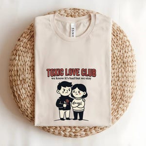 May include: Beige t-shirt with the text "TOXIC LOVE CLUB" in red and the phrase "we know it's bad but we stay". The shirt features a cartoon couple, one with a tear and the other with arms crossed.