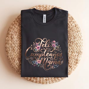 May include: Black t-shirt with a rose gold floral design and the words "Feliz Cumpleaños Mama". The design includes pink, purple, and blue flowers. The shirt is displayed on a woven, circular mat.