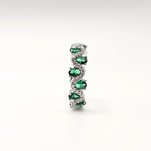 May include: A silver ring with a repeating pattern of oval emerald-green gemstones and smaller, clear, round-cut stones. The ring's design is set against a white background, accentuating the sparkle of the gems.