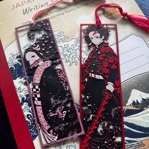 May include: Two rectangular metal bookmarks featuring anime characters. The left bookmark is pink with a black and pink character design. The right bookmark is red with a black and red character design. Both have matching tassels at the top.