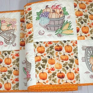 Puede incluir: Manteles individuales acolchados con temática otoñal con patrones naranjas y blancos. Los manteles individuales presentan imágenes bordadas de calabazas, una carretilla y un barril de madera lleno de productos. Los bordes están ribeteados con tela naranja.