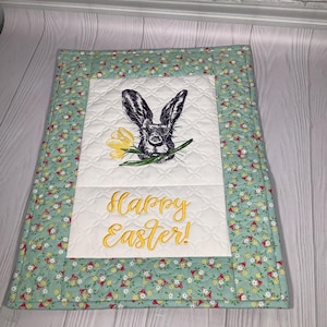 Może przedstawiać: Pikowana podkładka na stół z motywem wielkanocnym. Na środku czarno-biały królik trzymający żółty kwiat, z napisem "Happy Easter!" w kolorze żółtym. Obramowanie to kwiatowy wzór na jasnym zielonym tle.
