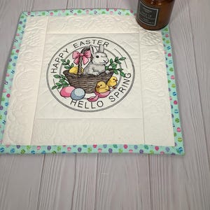 Może przedstawiać: Kwadratowa, pikowana podkładka pod talerz o tematyce wielkanocnej z haftowanym centralnym wzorem. Wzór przedstawia królika, pisklęta i pisanki w koszyku, ze słowami "Happy Easter Hello Spring". Podkładka ma jasnoniebieską obwódkę.