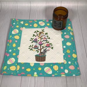 Może przedstawiać: Pikowana podkładka w tematyce wielkanocnej z turkusową obwódką z wzorami jajek. Środek ma haftowany wzór drzewka wielkanocnego z jajkami, królikiem i ptakiem. Na podkładce stoi brązowa świeca.