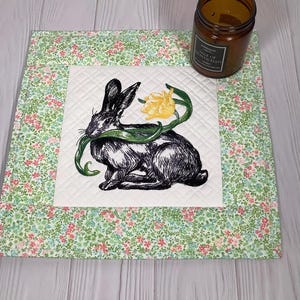 Może przedstawiać: Pikowana podkładka z kwiatowym wzorem i centralnym kwadratem z haftowanym czarnym królikiem z żółtym kwiatem. Na podkładce stoi brązowa szklana świeca.