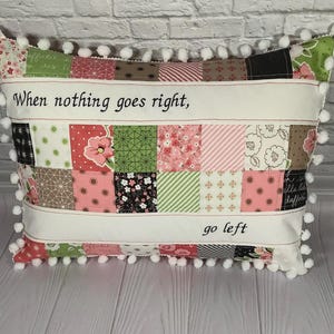 Könnte beinhalten: Ein rechteckiges Patchwork-Kissen mit verschiedenen floralen und geometrischen Mustern in Rosa, Grün und Braun. Das Kissen hat weiße Bänder mit dem Text "When nothing goes right, go left" und ist mit weißen Pompons verziert.