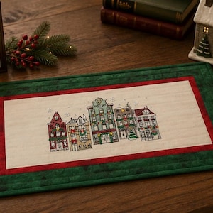 May include: A rectangular fabric placemat featuring a central cream-colored panel with an embroidered illustration of five multi-story buildings decorated for the holidays, framed by a red border and a wide, dark green quilted outer border.
