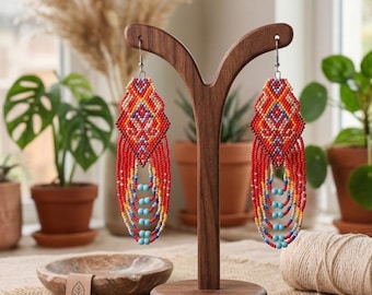 May include: A pair of beaded earrings in red, orange, yellow, and turquoise. The earrings are displayed on a wooden stand. The earrings have a diamond-shaped top and long, fringed bottoms. The background includes plants in terracotta pots.
