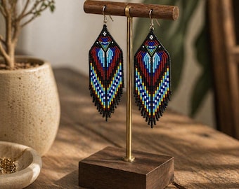 May include: A pair of beaded earrings displayed on a wooden stand. The earrings feature a geometric design with red, blue, yellow, and black beads. The earrings have a fringe at the bottom. The stand has a gold-colored metal rod.