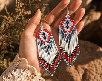 May include: A pair of long, beaded earrings with a diamond-shaped top in red, white, blue, and black. The earrings have a fringe of white beads with red and black accents. The earrings are held up in a hand.