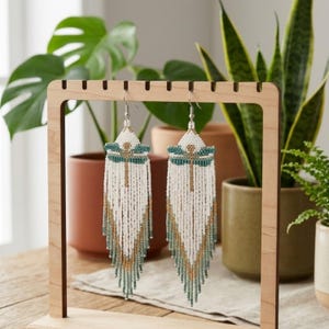 Pode incluir: Brincos de contas brancas com um design de libélula em verde-azulado e dourado. Os brincos têm uma longa franja e são exibidos em um suporte de brincos de madeira. Os brincos são de estilo boêmio.