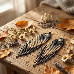 May include: A pair of beaded earrings with a geometric design. The earrings feature black, cream, and silver beads, with a fringe at the bottom. The earrings are displayed on a rustic wooden surface, surrounded by dried flowers and leaves.