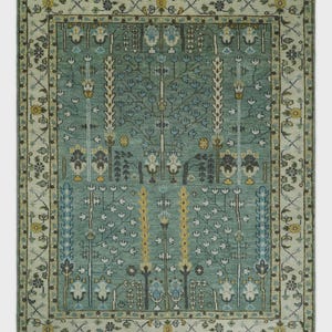 May include: A rectangular area rug with a teal background and an intricate floral design. The rug features a cream-colored border with floral and geometric patterns. The central design includes stylized trees and floral motifs in shades of blue, yellow, and gray.