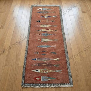 Puede incluir: Una alfombra rectangular de color salmón con un patrón repetido de peces estilizados en varios colores. Los diseños de peces incluyen detalles azules, naranjas y blancos. La alfombra tiene un borde oscuro y un borde con flecos.