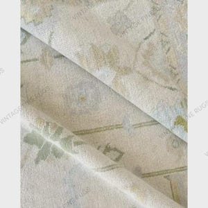 May include: Close-up of a folded, vintage rug with a floral pattern. The rug features a cream-colored background with faded green and blue floral designs. The text "VINTAGE FINE RUGS" is printed along the edges.