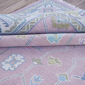 May include: Rolled-up area rug in shades of pink, blue, and green. The rug features a detailed floral pattern with a geometric border. The rug's design is visible on the rolled edges, showcasing its intricate details.