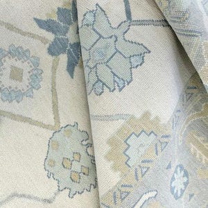 May include: Close-up of a rolled-up area rug with a floral pattern. The rug features a cream-colored background with blue, light green, and tan floral designs. The rug's texture is visible, and the edges are slightly curled.