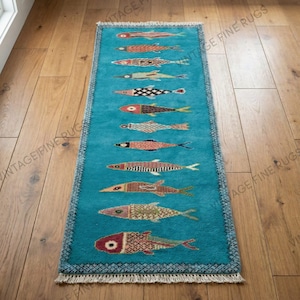 May include: A rectangular turquoise rug with a repeating pattern of colorful fish. The fish are in various colors and patterns, including red, orange, and black. The rug has a fringed edge and is placed on a wooden floor.