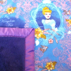 May include: A purple and blue fleece blanket with a satin border. The blanket features a print of Cinderella in a blue dress with a crown, surrounded by flowers and vines. The text "I'm in a Kingdom of my own" is printed on the blanket.