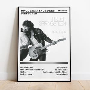 May include: Black and white framed poster featuring Bruce Springsteen with a guitar. The poster includes the text "BRUCE SPRINGSTEEN" and "BORN TO RUN" at the top, and a list of song titles at the bottom. The image is displayed on a wooden floor.