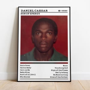 May include: A framed poster featuring a portrait of Daniel Caesar with the text "SON OF SPERGY." The poster has a red background and a list of song titles. The image is displayed on a white wall and a wooden floor.