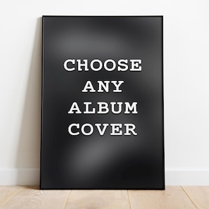 May include: A framed black poster with the text "CHOOSE ANY ALBUM COVER" in white, sans-serif font. The poster is leaning against a white wall on a wooden floor.