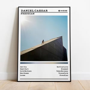 May include: A framed poster featuring the album "Freudian" by Daniel Caesar. The image shows a person walking on a building's angled roof against a blue sky. The poster includes the album title, artist name, and a tracklist.