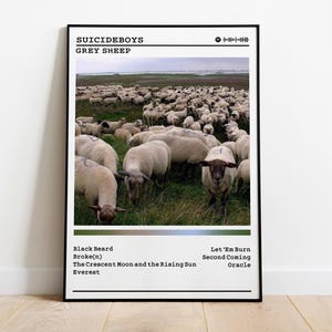 Puede incluir: Un póster enmarcado con una fotografía de un rebaño de ovejas pastando en un campo. El póster incluye el texto "SUICIDEBOYS GREY SHEEP" en la parte superior, con una lista de títulos de canciones debajo. La imagen está colocada contra una pared blanca y un suelo de madera.