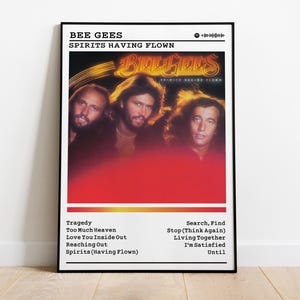 Puede incluir: Un póster enmarcado del álbum "Spirits Having Flown" de los Bee Gees. La imagen muestra a los miembros de la banda con un fondo degradado rojo y amarillo. El póster incluye el título del álbum, el nombre de la banda y una lista de canciones.