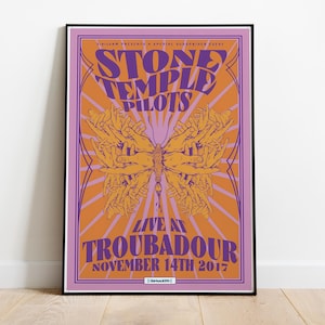 Puede incluir: Un póster enmarcado del concierto de Stone Temple Pilots en el Troubadour el 14 de noviembre de 2017. El diseño presenta una mariposa hecha de manos, con texto en tonos morados y naranjas. El póster está en un marco negro.