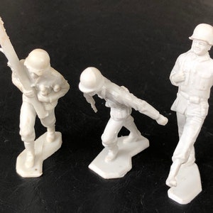 May include: Three white plastic toy soldiers. One soldier is holding a bazooka, one is holding a rifle, and one is standing at attention.