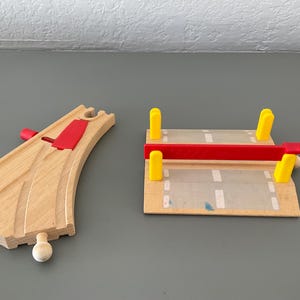 May include: Wooden toy train track pieces. One curved track with a red switch and a bridge with yellow supports and a red barrier. The bridge has a road design.
