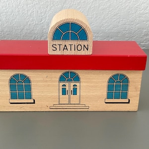 May include: Wooden toy train station with a red roof and light wood walls. The station has blue arched windows and doors. The word "STATION" is printed on the top of the building.