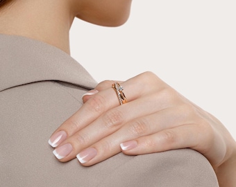 May include: A gold-toned ring with a small, clear gemstone and smaller accent stones. The ring is on a person's finger, with a neutral-toned background. The ring's design features a crisscross band and is likely a piece of jewelry.
