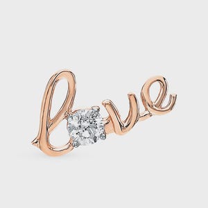 Könnte beinhalten: Roségoldfarbener Schmuck, der das Wort „love“ schreibt, mit einem funkelnden, runden, klaren Edelstein in der Mitte. Die elegante Schrift ist kursiv und vor einem schlichten Hintergrund.