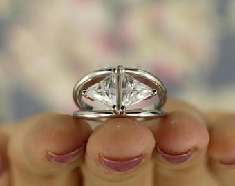 Pode incluir: Um anel de prata com duas pedras preciosas claras, lapidadas em forma de triângulo, colocadas lado a lado. O anel tem um design único com uma banda dupla e um elemento decorativo que liga as pedras. O fundo é suave e desfocado.