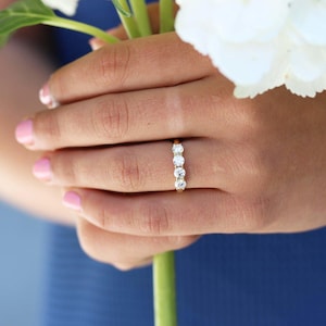Può includere: Un anello in oro con quattro pietre preziose rotonde e trasparenti disposte in fila. L'anello è su un dito, con uno sfondo blu e un fiore bianco sullo sfondo.