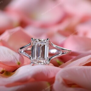 Op de afbeelding: Een zilveren ring met een grote, rechthoekige, heldere edelsteen. De ring heeft een split-schachtontwerp, waarbij de band zich splitst in twee delicate armen aan weerszijden van de edelsteen. De ring is geplaatst tegen een zachte roze achtergrond.