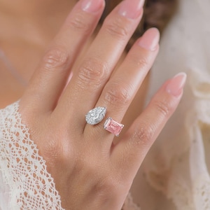 Puede incluir: Un anillo de plata con dos piedras preciosas: una piedra clara en forma de pera y una piedra rosa rectangular. El anillo se muestra en una mano con uñas cuidadas, destacando el diseño de la joya. El diseño abierto permite que las piedras preciosas se asienten en dedos adyacentes.