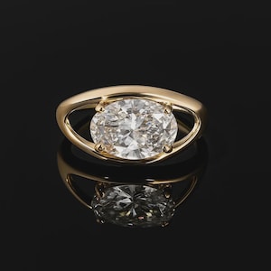 May include: A gold ring with a large, oval-cut diamond. The diamond is set in a bezel setting, and the band has a unique, open design. The ring is displayed on a black surface, with its reflection visible below.