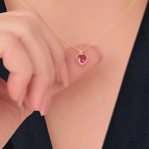 Puede incluir: Un collar dorado con un colgante en forma de corazón. El colgante presenta una piedra preciosa roja brillante engastada en un marco dorado. El collar es delicado y se lleva alrededor del cuello.