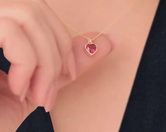 May include: A gold-colored necklace with a heart-shaped pendant. The pendant features a vibrant red gemstone set within a gold frame. The necklace is delicate and appears to be worn around the neck.