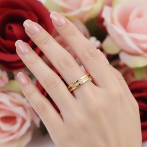 Puede incluir: Un anillo dorado con un diseño de cruz, engastado con pequeñas piedras preciosas transparentes. El anillo está en un dedo, con un fondo borroso de rosas rojas y rosas.