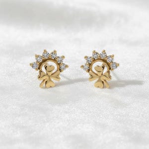 Könnte beinhalten: Ein Paar goldfarbene Ohrstecker. Jeder Ohrring hat ein Kleeblatt-Design mit einem herzförmigen Mittelteil und einem Kranz aus kleinen, klaren Edelsteinen. Die Ohrringe sind vor einem weißen Hintergrund platziert.