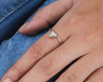 May include: A gold ring with a triangular, clear gemstone. The ring is simple and elegant, with a thin band and a geometric design. The ring is worn on a finger, against a background of denim.