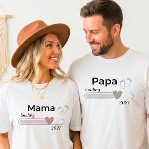 May include: Two white short-sleeved t-shirts. The left shirt features the text Mama loading, a pink heart, baby footprints, and the year 2027. The right shirt features the text Papa loading, a grey heart, baby footprints, and the year 2027.